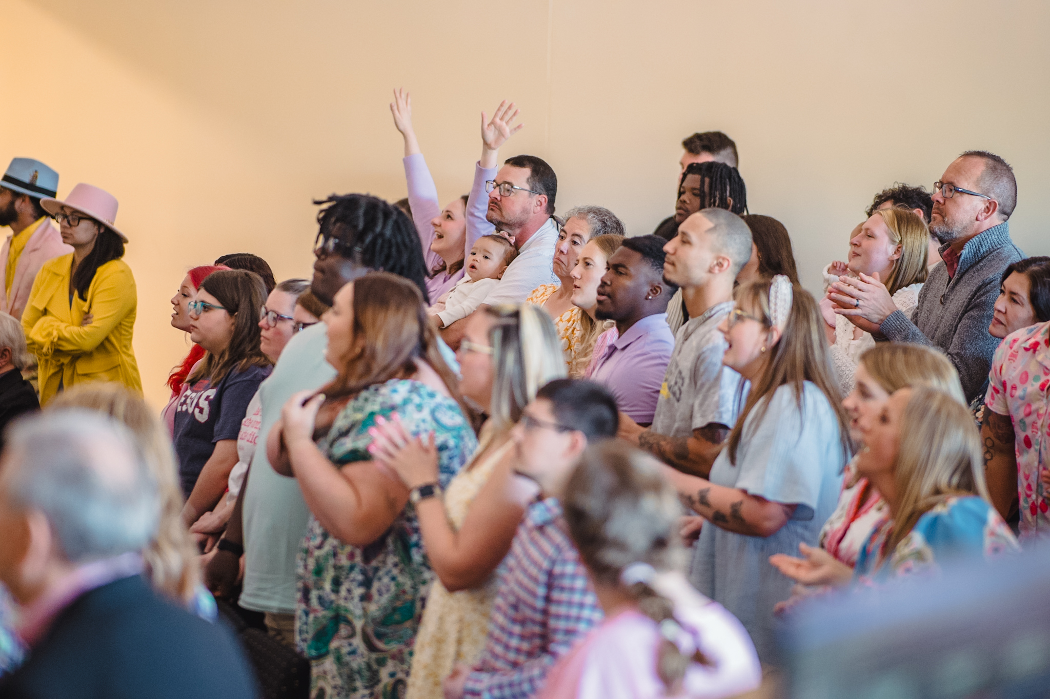 Diverse crowd of people standing and clapping at a large indoor event, several hands raised in the air.