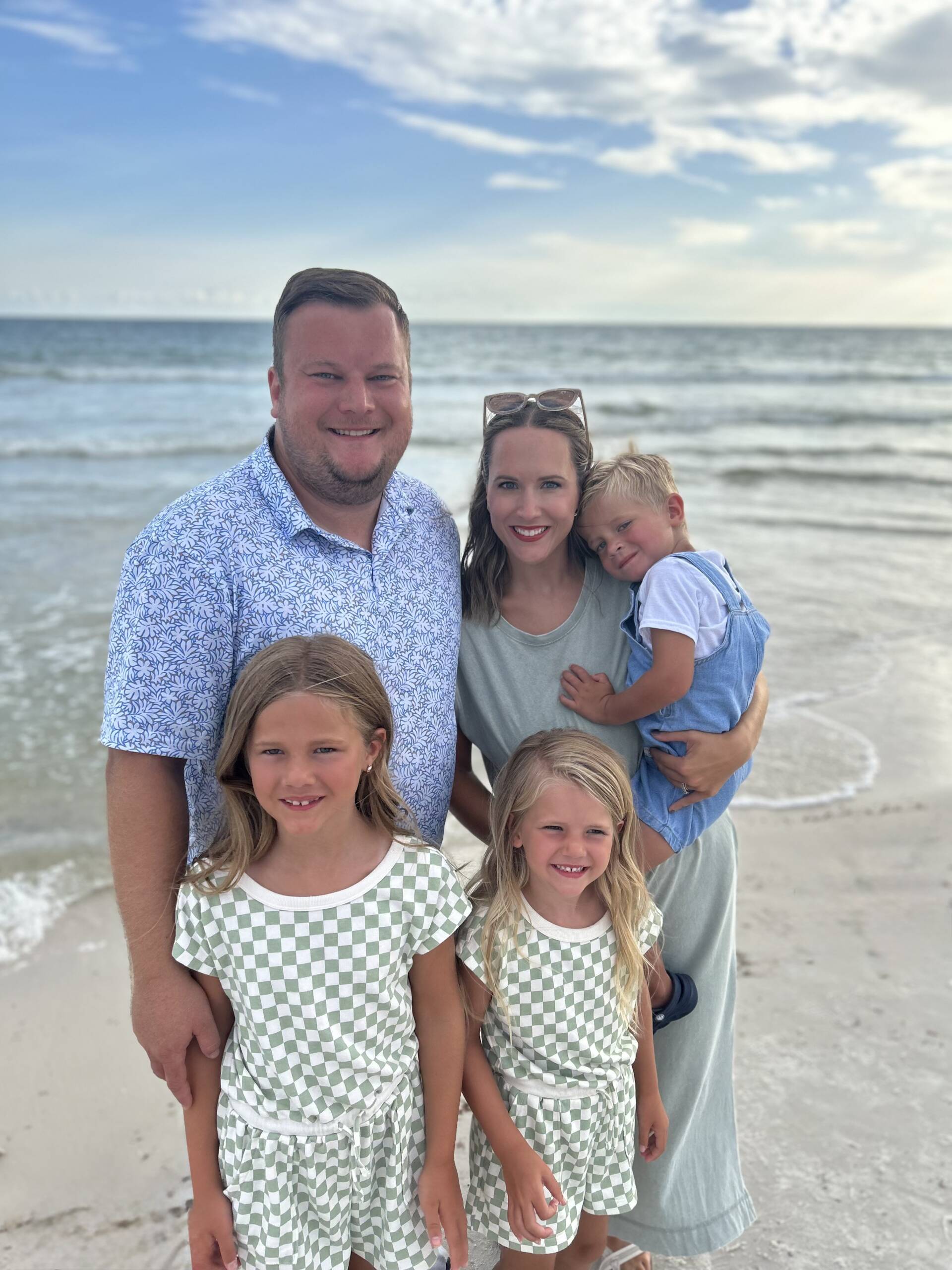 Smiling family of five posing on the beach with the ocean in the background; man in a blue patterned shirt, woman in green top, two girls in matching green-and-white checked dresses, and a boy in blue overalls.