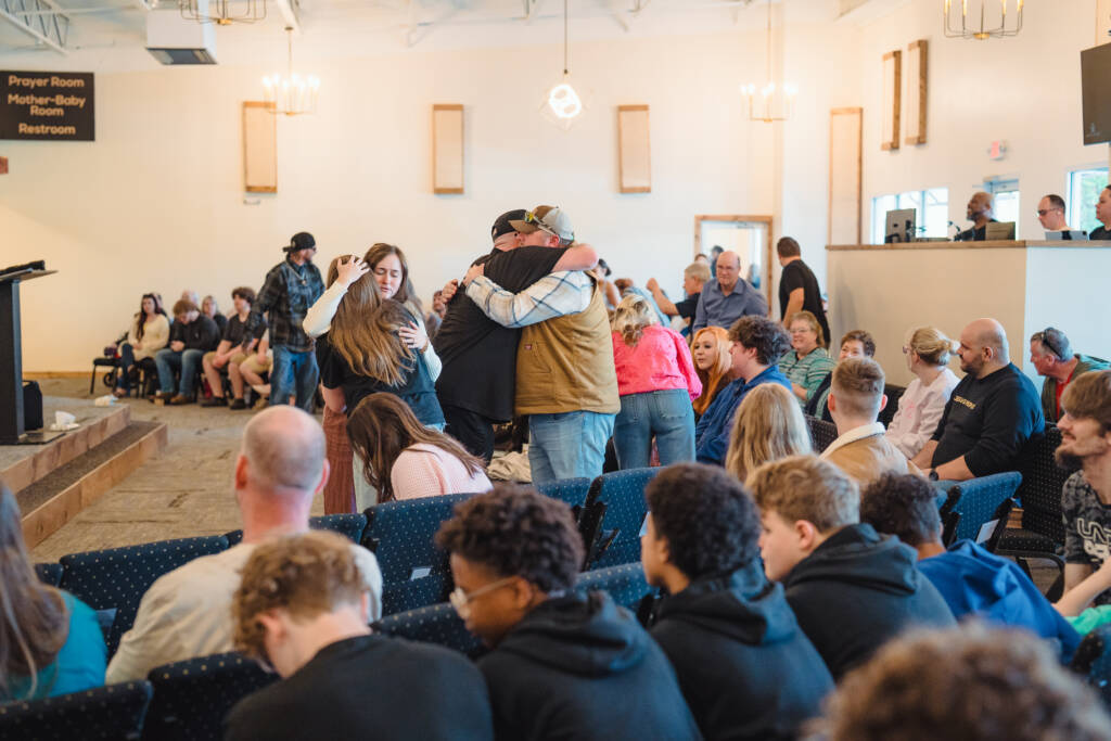 Group of people hugging in a crowded meeting hall, with others seated and watching in the background.