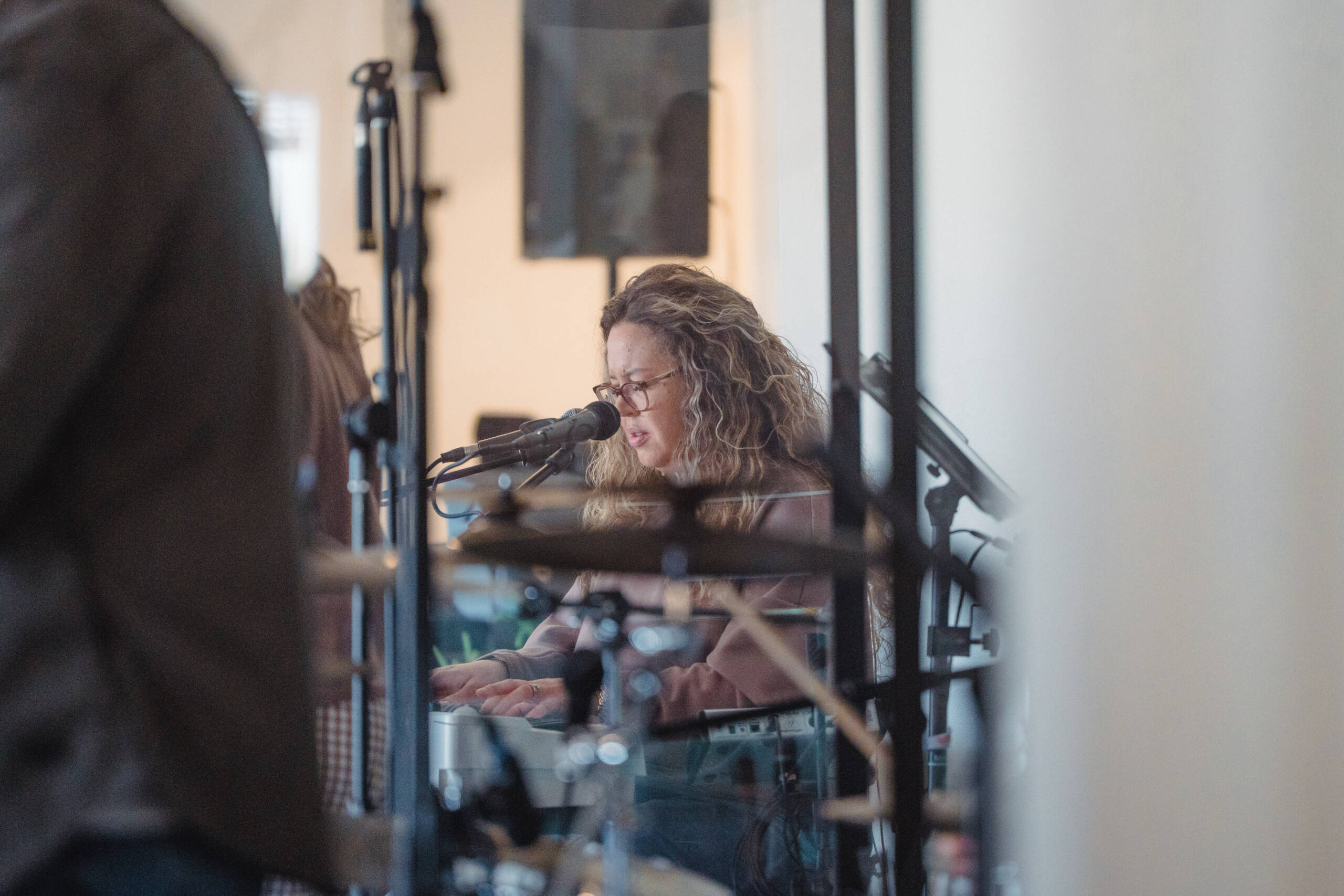 Musician with curly hair and glasses singing into a microphone in a recording studio, seen through glass panels with equipment in the foreground